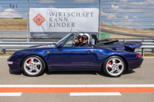 Blauer Porsche vor Wirtschaft kann Kinder e. V. Banner auf der Rennstrecke
